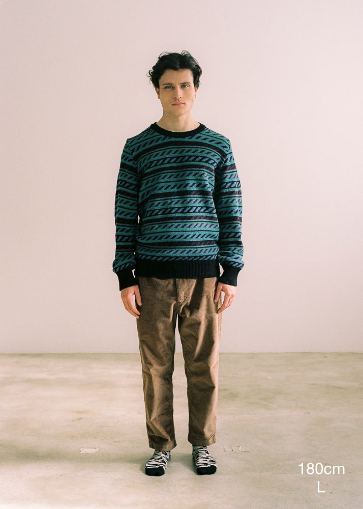 Fotografía frontal de cuerpo entero en la que se detalla altura del modelo (180cm) y la talla de jersey (M). El modelo lleva el jersey unisex IAIOS con cenefa en tonos azul, verde y negro, moda sostenible hecha en Catalunya (España) con hilo reciclado, ideal para invierno.