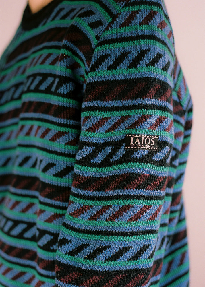 Fotografía de detalle de la etiqueta con el logo de IAIOS en la parte externa de la manga. Jersey unisex IAIOS con cenefa en azul, verde y negro, detalle del tejido, moda sostenible elaborada con hilo reciclado en talleres de Catalunya (España).