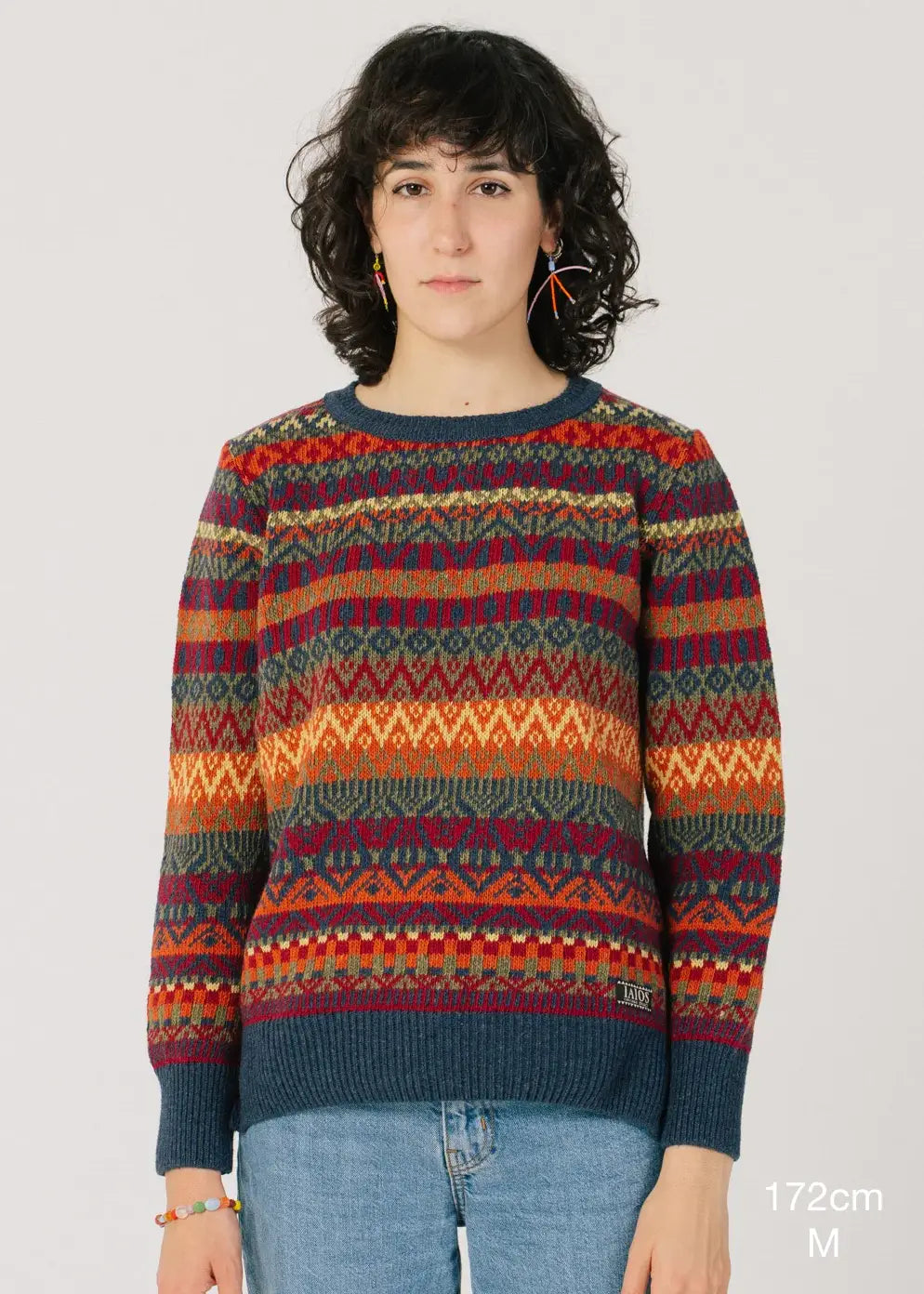 Fotografía de cuerpo entero en la que se detalla la altura de la modelo (172cm) y la talla de su jersey (M). El modelo lleva el jersey IAIOS unisex colorido navideño con diseño nórdico de cinco colores, moda sostenible elaborada en talleres de Catalunya (España) con hilo reciclado.