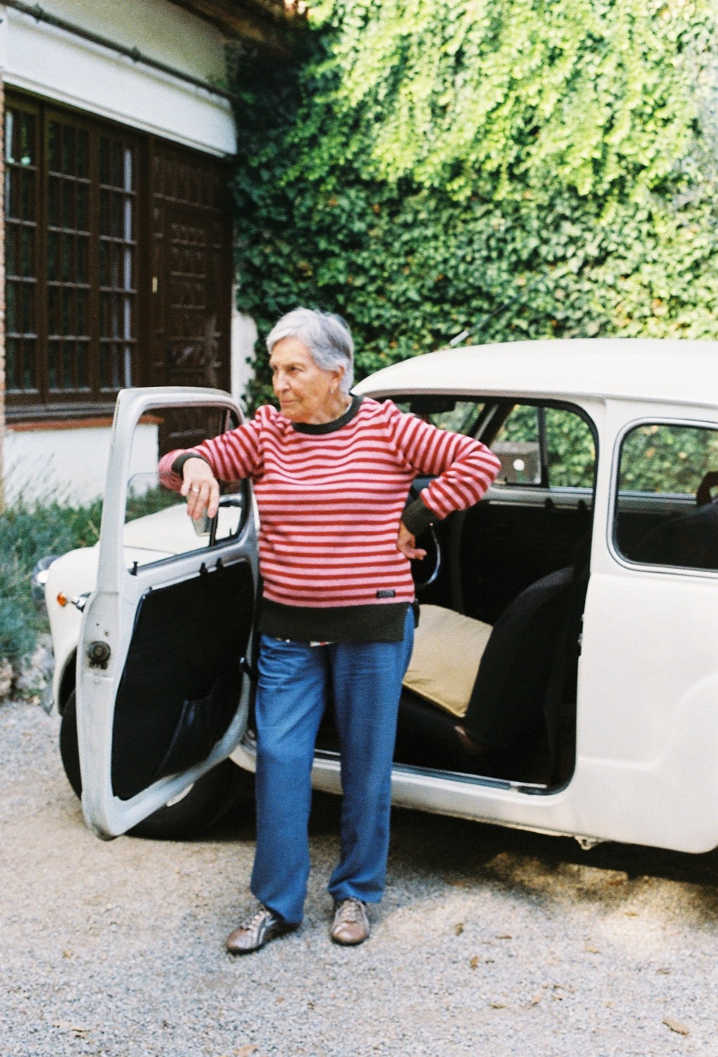 La abuela apoyada a su coche lleva el jersey unisex IAIOS Truth de rayas rojas y rosas con puños y cuello verde oscuro, moda sostenible hecha en Catalunya (España) con hilo reciclado.