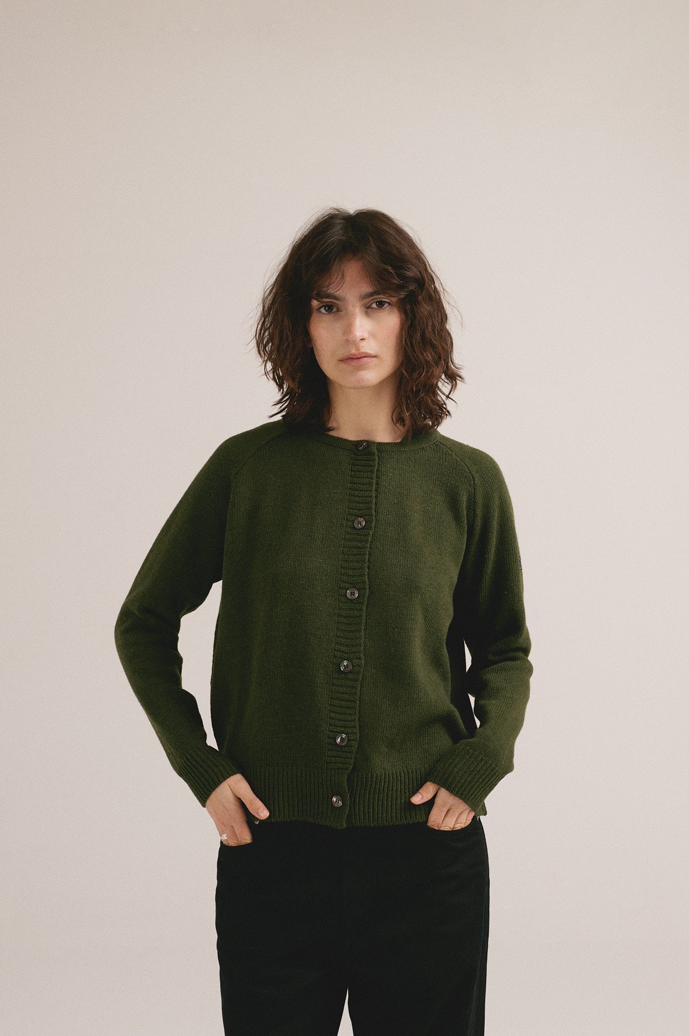 Mujer con cárdigan de punto verde con botones de IAIOS y pantalón oscuro en fondo neutro.