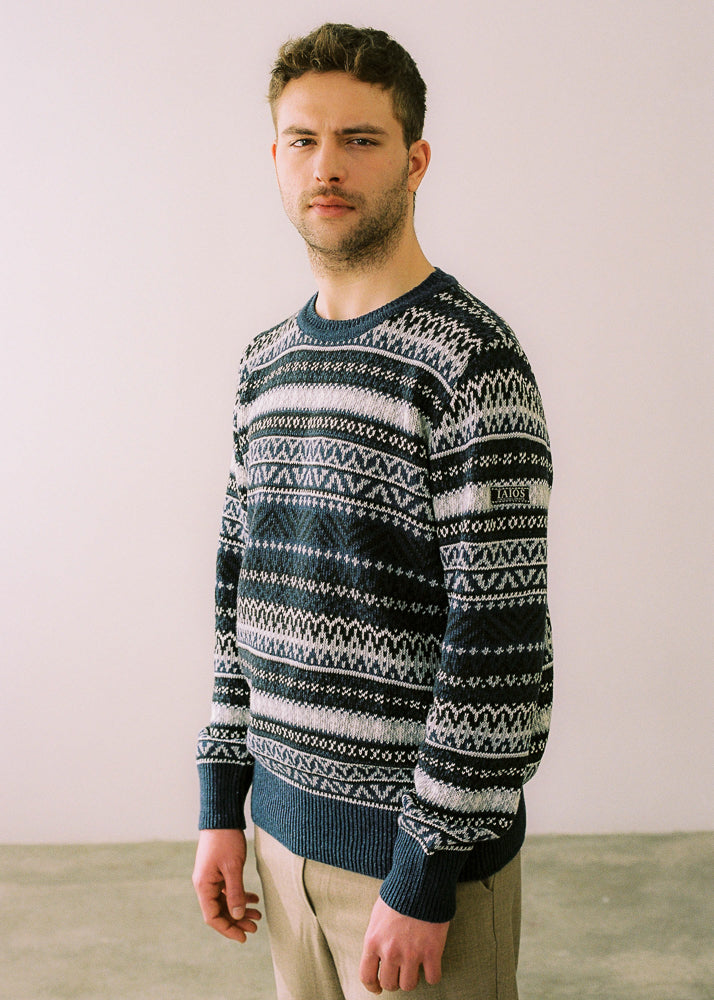 Fotografía de medio cuerpo, visión lateral en la que se aprecia la etiqueta con logo de IAIOS en la manga del jersey. El modelo lleva el jersey IAIOS unisex azul estilo nórdico, tejido jacquard de cinco colores, moda sostenible elaborada en talleres de Catalunya (España) con hilo reciclado.