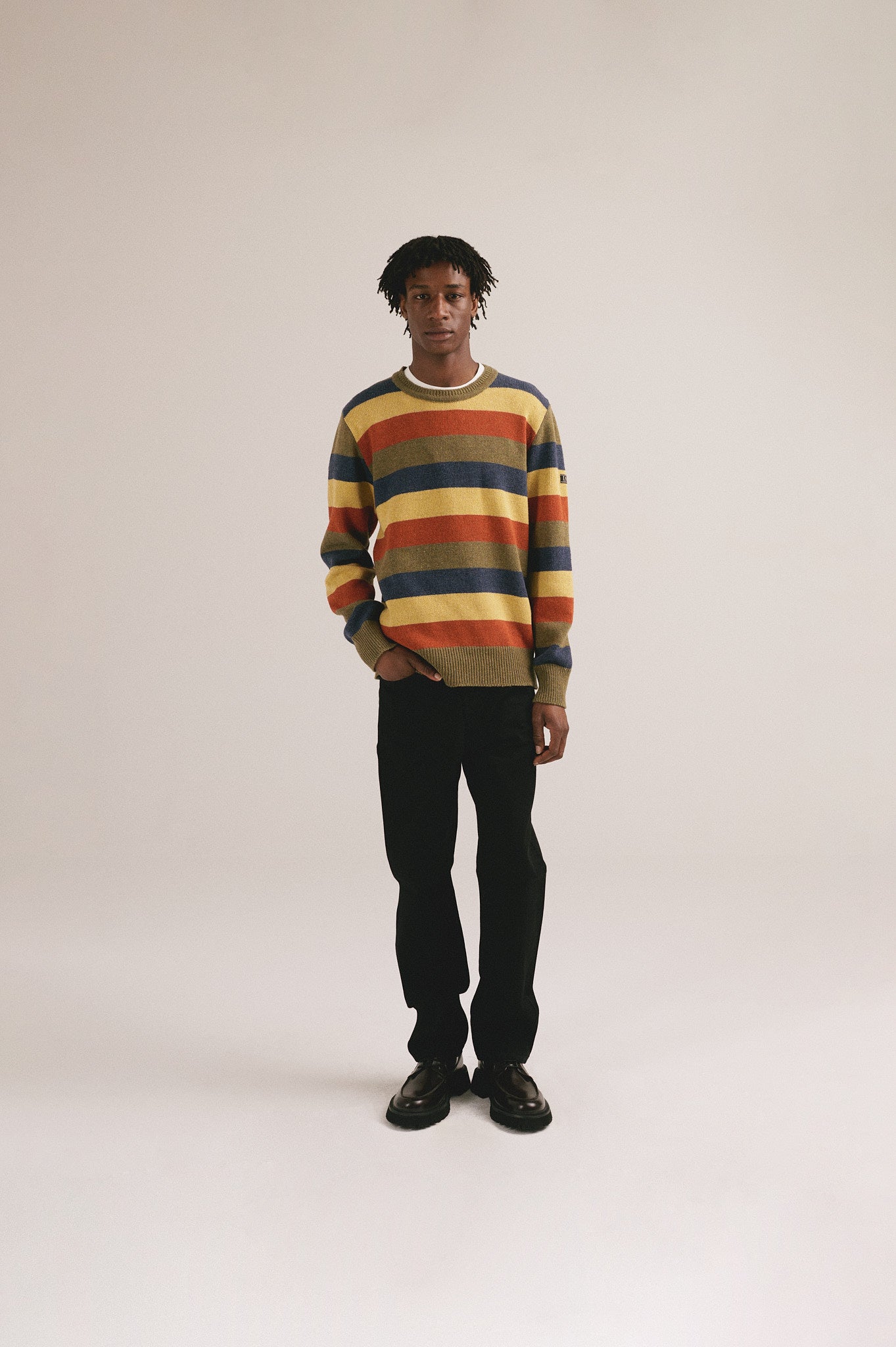 Hombre con jersey de punto a rayas multicolor de IAIOS en tonos amarillo, azul y naranja y pantalón oscuro.