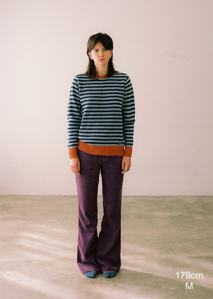 Fotografía frontal de cuerpo entero donde se detalla la altura de la modelo (179cm) y la talla del jersey (M). La modelo lleva el jersey IAIOS unisex de rayas azul y naranja, moda sostenible elaborada en talleres de Catalunya (España) con hilo reciclado.
