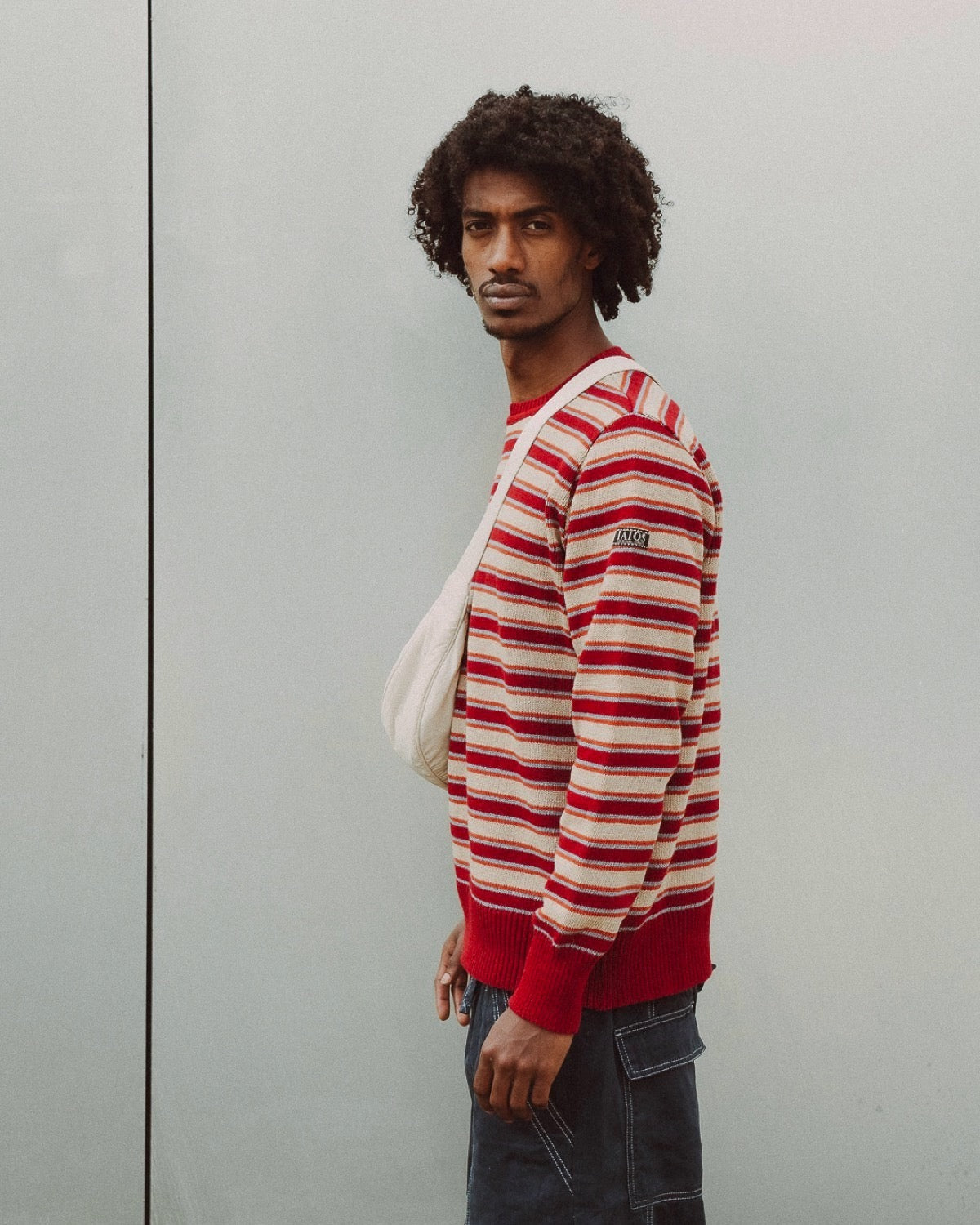El modelo lleva el jersey unisex IAIOS de rayas en tonos cálidos con predominio del rojo, combinado con beige y naranja, cuello redondo, hecho en Catalunya (España) con hilo reciclado.