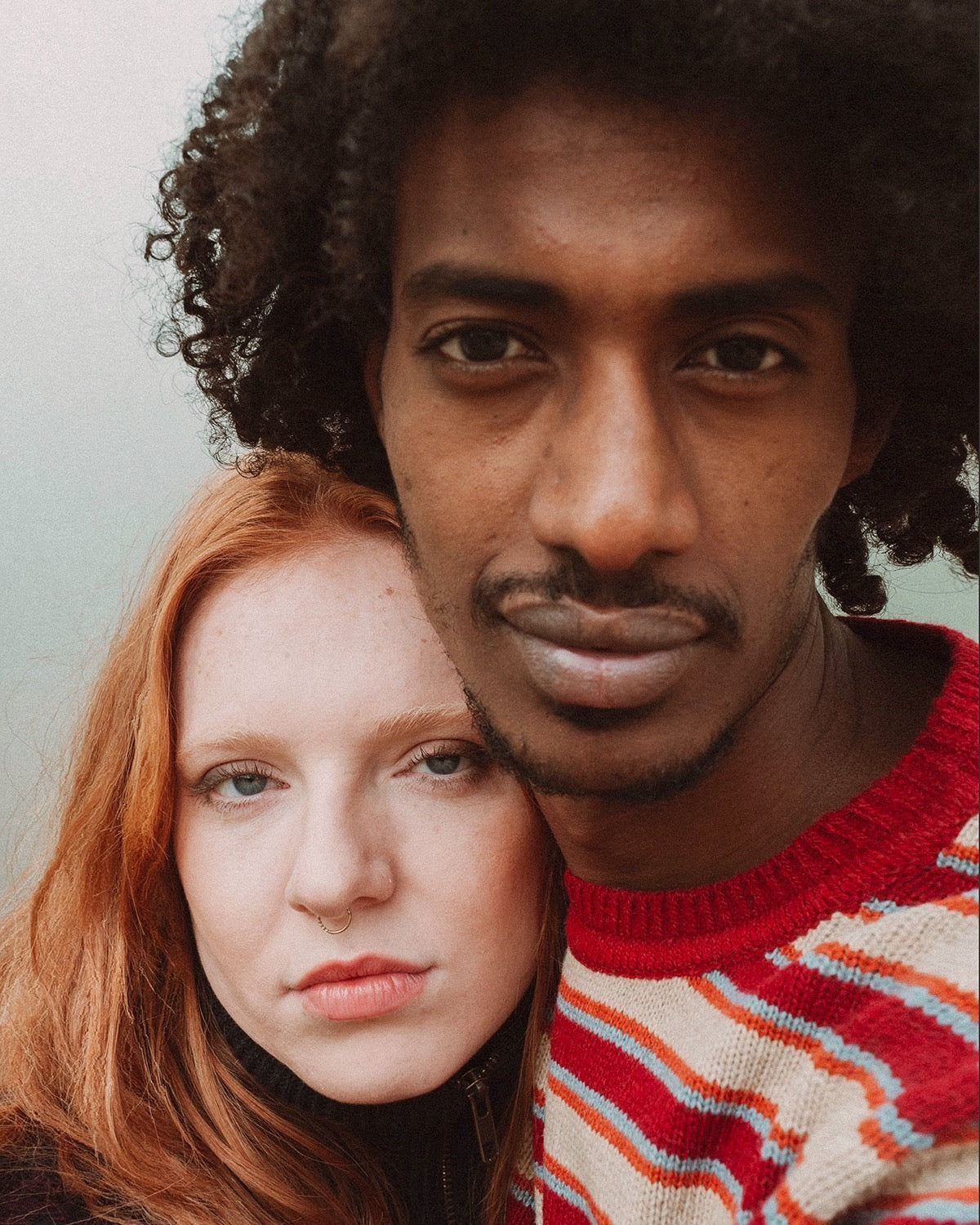 retrato de los modelos. Él lleva el jersey IAIOS unisex de rayas rojas, beige y naranjas, con cuello redondo y ajuste clásico, tejido con hilo reciclado en talleres de Catalunya (España).