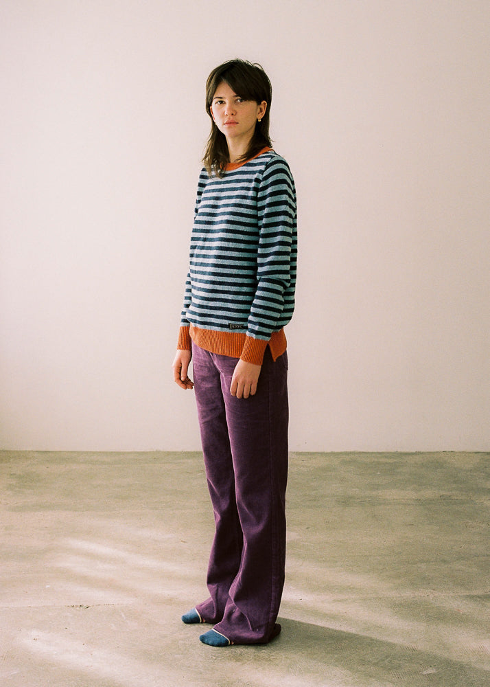Fotografía de cuerpo entero, se aprecia el jersey en visión lateral, combinado con pantalón de color lila. Jersey unisex IAIOS de rayas azul y naranja, moda sostenible hecha en Catalunya (España) con hilo reciclado, perfecto como regalo de Navidad.