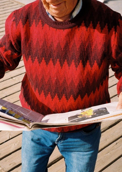 Fotografía escenificada, persona mayor leyendo el diario llevando este jersey color rojo y granate.